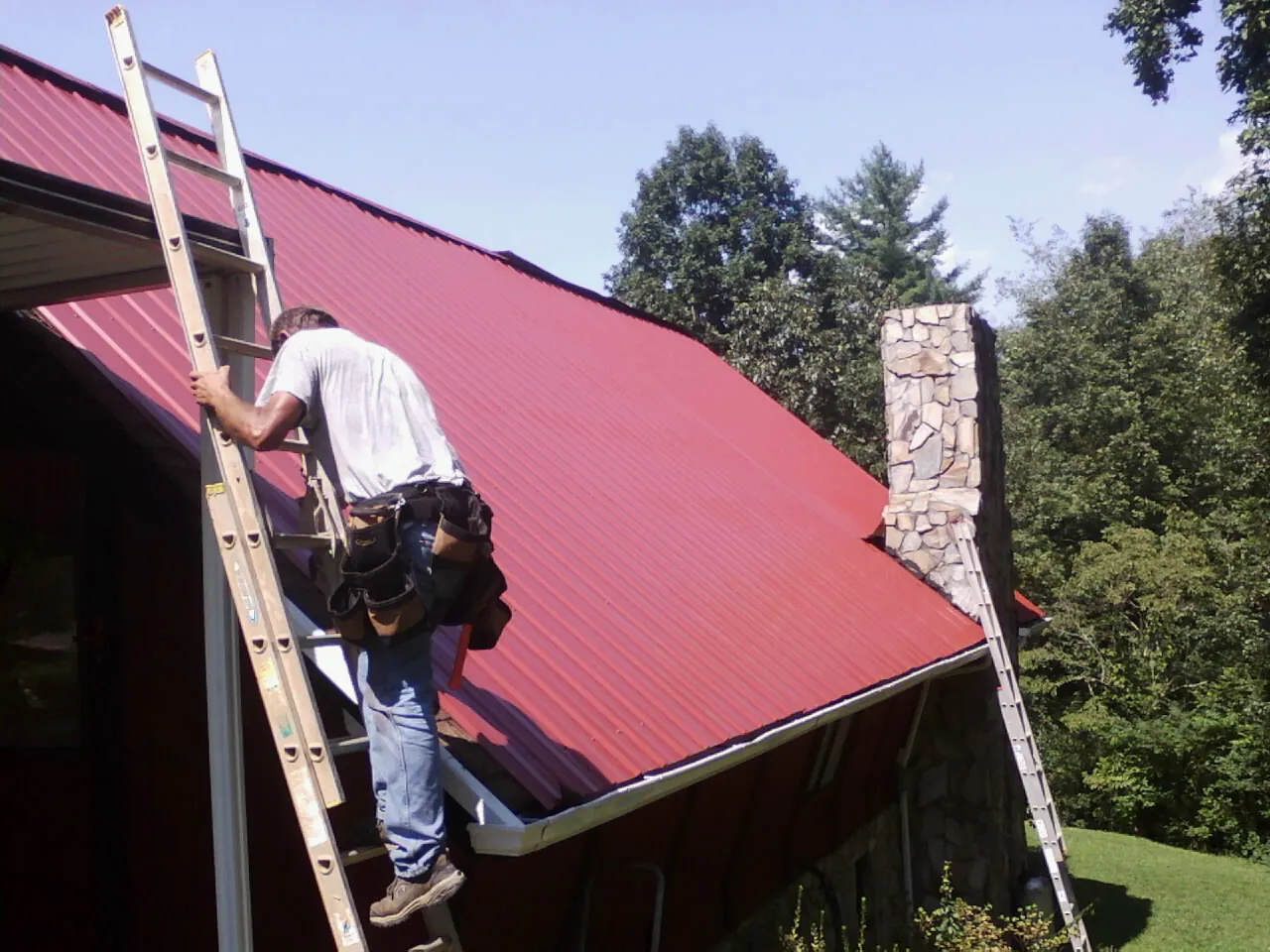 Roofer on a ladder beside a red metal roof and stone chimney in Richmond Hill